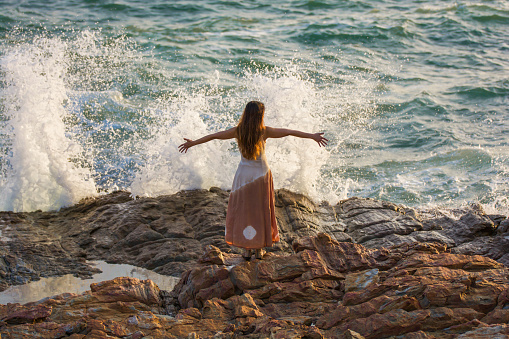 The wave is breaking on the stone. Beautiful Asian woman standing on the stones nearly waves splash with sea and sky background. Scenic View Beach Against Sky During Sunset.