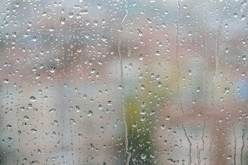 raining, raindrops on windowpane over blurred city view. Rain droplets on window against blur background  or surface with buildings or houses while raining with selective focus and copy space
