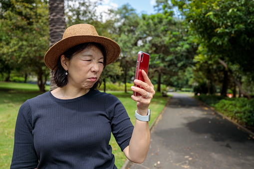 Asian woman with a hat in a park on a sunny day holding her phone with a troubled expression.