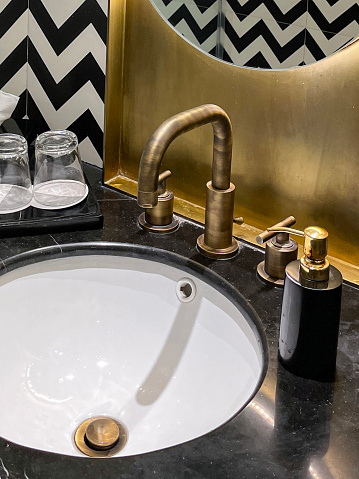 Stock photo showing close-up view of a luxury hotel bathroom with black and white zig-zag patterned wall tiles. Folded white towels hung on towel rail on sink unit with dual knob monobloc mixer tap in front of mirror.