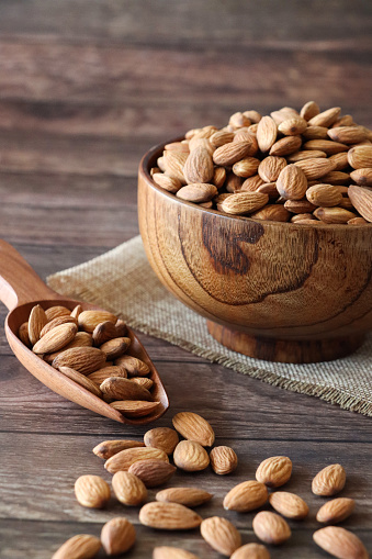 Stock photo showing close-up, elevated view of some almonds that are piled high in a wooden dish, against a woodgrain background. Raw almonds are considered to be a very healthy snack food, containing vitamin E, antioxidants and protein, and boasting a list of health benefits while aiding blood sugar control.