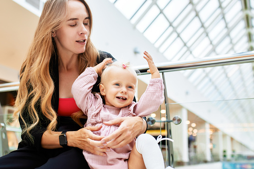 Young pretty mother with a small baby sitting on a bench in a shopping center. Mom and daughter in pink clothes hug, play, relax and have fun while shopping. Family weekend in shopping mall.