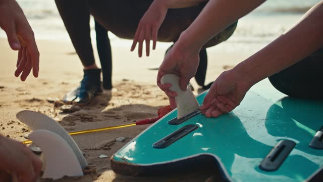 Close-up shot of a male surfer repairing his green surfboard and disassembling its fins. A man disassembles the surf with his own hands on the Sandy seashore
