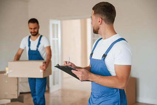 Writing in notepad, holding box. Two moving service employees in a room.