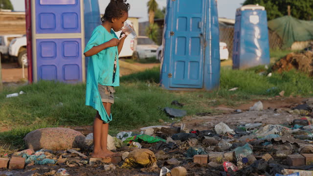 Poverty , poor, Black African child brushing her teeth next to portable toilets and stinking effluent water in an informal settlement