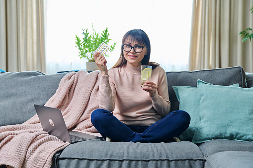 Middle-aged woman taking dietary supplement, fish oil capsule, omega 3. Mature female in home interior, sitting on sofa, diet, proper nutrition, medicine, health care, food supplements and people