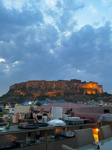 Jodhpur, Rajasthan, India - April, 11 2023: Stock photo showing people sitting eating at rooftop restaurant with view of Jodhpur's Mehrangarh Fort and Old Quarter rooftop cityscape. The buildings in the city's Old Quarter are painted an almost uniform blue, as to why? Nobody can agree on a reason.