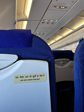 Stock photo showing close-up view of the interior of an airplane with rows of seats and in an economy class cabin of a passenger plane. A seat is viewed from the rear with a built-in fold down table displaying an information label informing passengers of the location of life vest safety equipment.