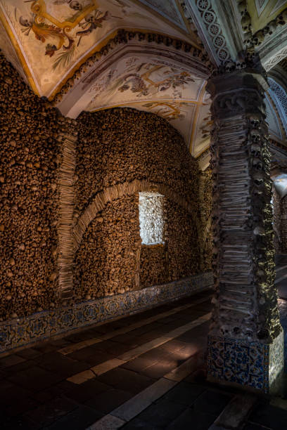 Column covered and decorated with human bones and skulls and the vaulted ceiling with religious allegorical paintings of The Chapel of Bones illuminated with soft light. Evora, Portugal.