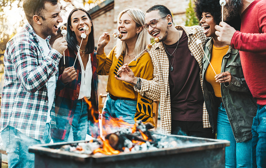 Happy friends roasting marshmallows over campfire - Group of young people enjoying garden party at summer vacation - Friendship concept with guys and girls having fun hanging outside together