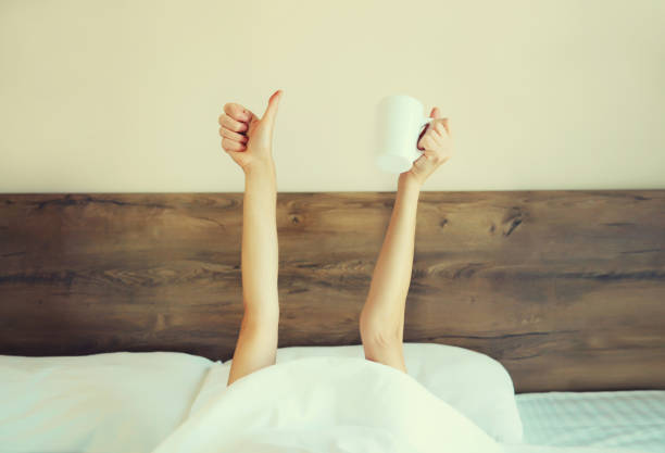 istock Cheerful lazy woman waking up after sleeping lying in soft comfortable bed showing empty cup coffee stretching her hands up from under the blanket in bedroom at home 1834784464