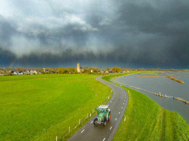 Tractor driving on a levee along the flooded river IJssel near Zalk village in Overijssel with storm clouds and a rainbow in the background. The floodplains are overflown after a long period of heavy rain upstream during autumn.
