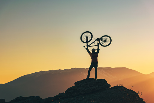 Happy biker stands in winner success pose with mtb bike above his head at mountain top and looks at sunset