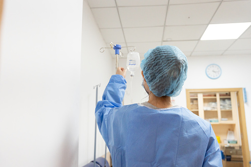 This is a photo of a medical professional in a hospital setting. The photo shows a medical professional wearing a blue surgical gown and blue hairnet adjusting an IV drip hanging from a metal pole. The scene might be performing some medical procedure or providing care to a patient.
