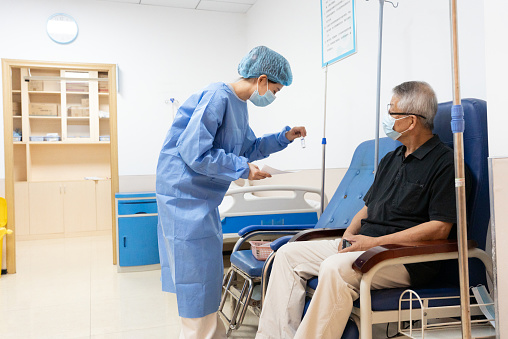 This photo shows a health care worker vaccinating a patient against COVID-19 at a clinic. The medical staff wore blue surgical gowns, hairnets, and masks on their faces. The patient sits on a blue chair with an IV pole next to him. The background is a white wall and a cabinet with a clock hanging on it.