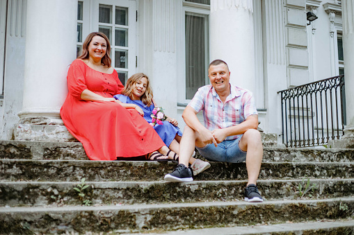Beautiful woman is sitting on steps of wide staircase next to her charming daughter and husband.