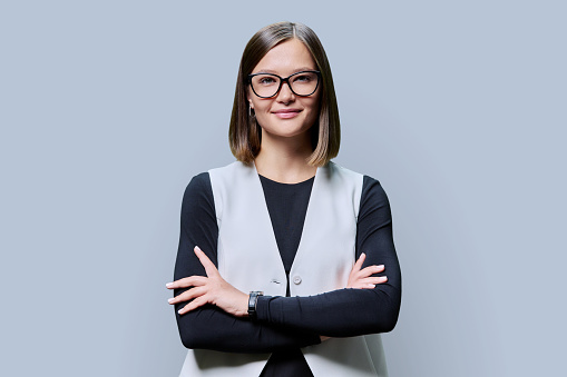 Young confident trendy smiling woman in eyeglasses with crossed arms looking at camera on gray studio background. Beauty, business, work entrepreneurship business expert owner agent concept