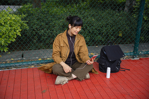 A woman sitting on the court watching a phone and listening to music