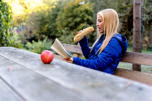 Tucked away on a bench, a young woman delves into a book while relishing the flavors of a sandwich, merging culinary and literary pleasures in nature's embrace