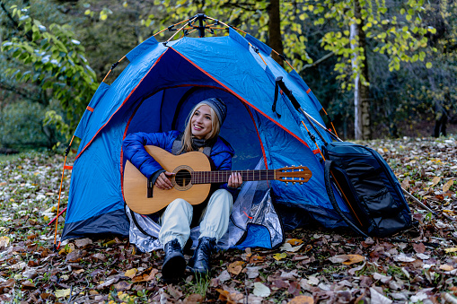 Experience a peaceful scene as a young woman sits in front of her tent, strumming the guitar, creating a harmonious melody that resonates with the tranquility of the natural surroundings