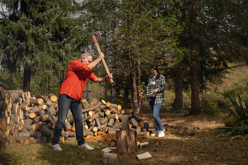 Lumberjack cutting firewood with axe outdoors in nature, his wife is helping him.