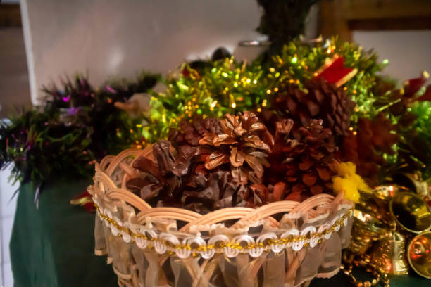 Captured in the soft luminescence of a quiet afternoon, this evocative image presents a rustic wicker basket, carefully positioned on an antique wooden table that speaks of age and elegance. It is generously filled with a collection of pine cones, each one a delicate treasure from the whispering pines standing sentinel in the distant woods. The textures are rich and compelling, ranging from the woven threads of the basket to the wooden grooves on the table and the intricate patterns of the pine cone scales. The ambiance is one of serenity and warmth, with the natural light accentuating the depth of the forest’s bounty. This stunning still life is not just a picture; it's an invitation to embrace the stillness and beauty of nature's own artistry
