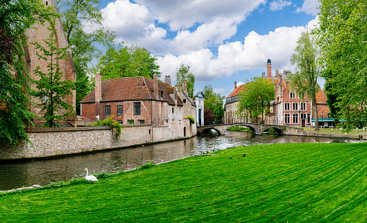 View of a canal in Bruges, Belgium. The canal is lined with historic brick buildings and a stone bridge. In the background, a church spire can be seen rising above the rooftops. The photo was taken on a sunny day, with blue skies and green trees visible.