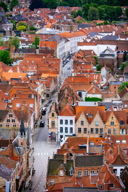 An aerial view of a Bruges town with red roofs. The buildings are tightly packed together and the streets are narrow. The photo is taken from a high angle, looking down on the town. The buildings are mostly made of brick and have a variety of architectural styles. The roofs are mostly red and orange, with some gray and white mixed in. The photo is taken during the day, with the sun shining on the buildings.