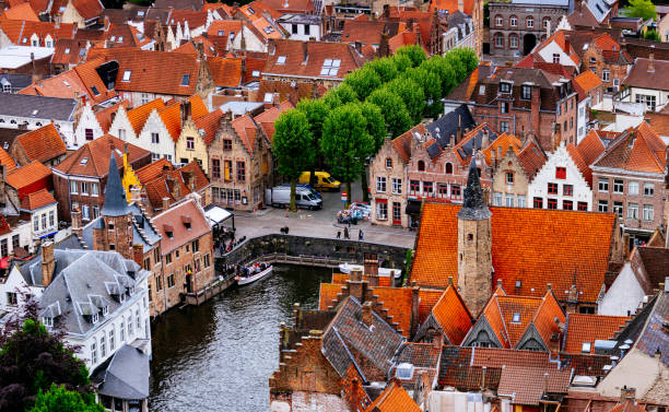 An aerial view of a Bruges town with red roofs. The buildings are tightly packed together and the streets are narrow. The photo is taken from a high angle, looking down on the town. The buildings are mostly made of brick and have a variety of architectural styles. The roofs are mostly red and orange, with some gray and white mixed in. The photo is taken during the day, with the sun shining on the buildings.