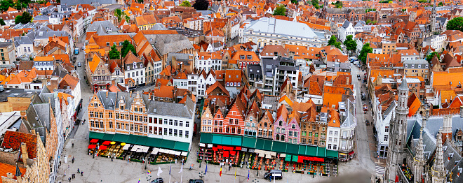 An aerial view of the historic city of Bruges and Grote Markt, Belgium. The photo captures the city’s medieval architecture, with its red brick buildings and orange tiled roofs. The photo also shows the city’s narrow streets and canals. The photo was taken from a high vantage point, giving a bird’s eye view of the city.