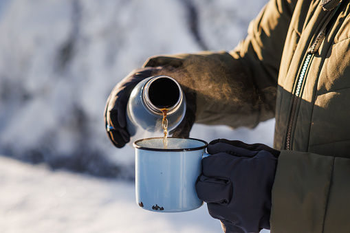 Hiker pouring hot drink from thermos into travel mug. Refreshment during winter trekking in cold weather