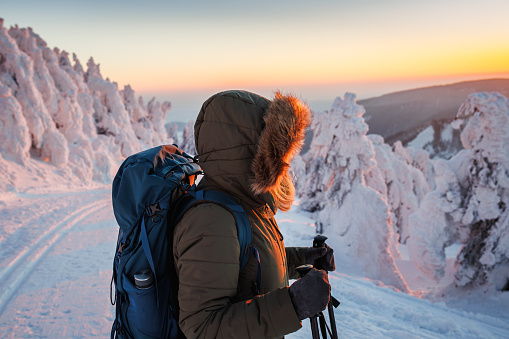 Hiking in winter mountains during sunset. Woman with backpack wearing winter coat with hood. Sports warm clothing for outdoor activity