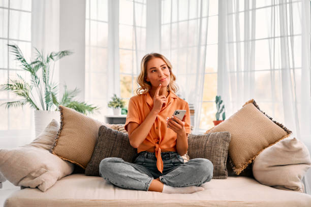 Attractive woman touching chin and looking thoughtfully aside while sitting on sofa with cell phone. Technology and lifestyle. Blonde female with crossed legs resting at home and using modern gadget. day dreaming stock pictures, royalty-free photos & images