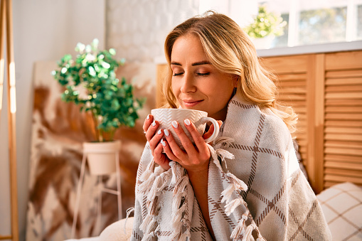 Dreamy caucasian woman covered with checkered blanket warming with hot tea while sitting on bed during autumn season at cozy bright apartment.