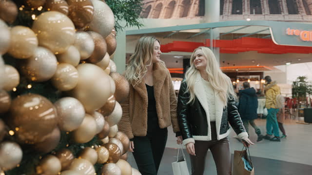 SLO MO Cheerful Young Female Friends in Trendy Clothes With Paper Bags Talking While Walking Along Christmas Tree in Shopping Mall