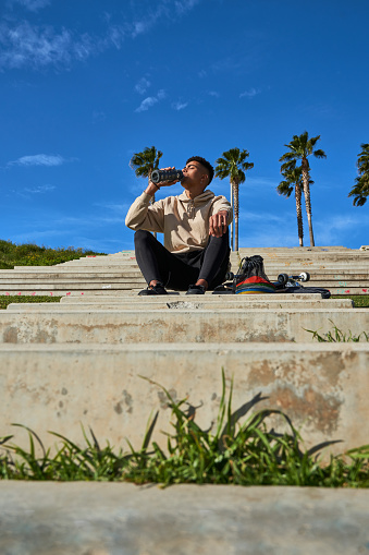 Low angle full body of Hispanic guy sitting on stone steps and drinking water for refreshing after training on skateboard in sunny urban park
