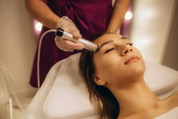 Side view portrait of a beautiful female brunette leaning on a bed with closed eyes while having rejuvenation electroporation on her face by a cosmetologist. High quality photo