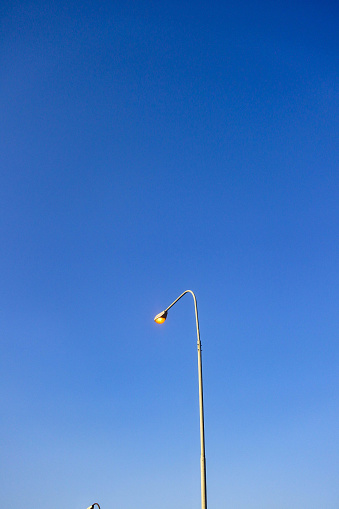 As the blue hues of dusk settle in, the solitary street light stands as a beacon of guidance on the side of a meandering road. This image captures the essence of transition from day to night, where the light post becomes a luminary against the creeping shadows. The warm glow of the lamp brings a sense of calm and safety, its light a soft halo in the encroaching twilight. The road, bordered by the subtle shapes of the natural landscape, leads into the unknown, and the street light, with its steadfast presence, promises continuity in the journey ahead. This scene perfectly encapsulates the juxtaposition of man-made structure and nature's expansiveness, suitable for themes of travel, solitude, and the passage of time. The photograph mesmerizes its audience, inviting them to consider the quiet moments of beauty that often go unnoticed in the rush of daily life