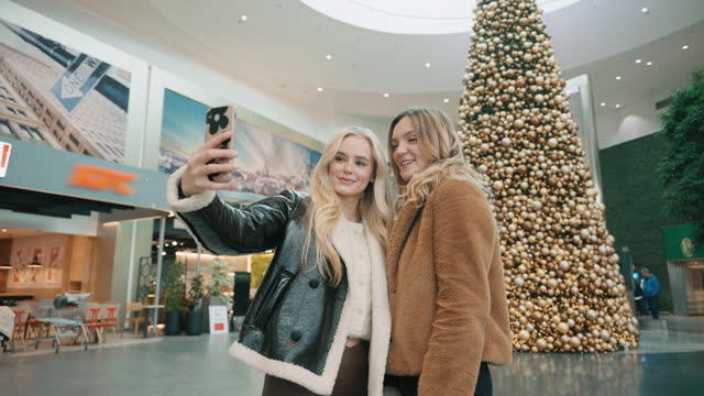 SLO MO Beautiful Young Female Friends in Trendy Clothes Taking Selfie With Huge Christmas Tree in Shopping Mall