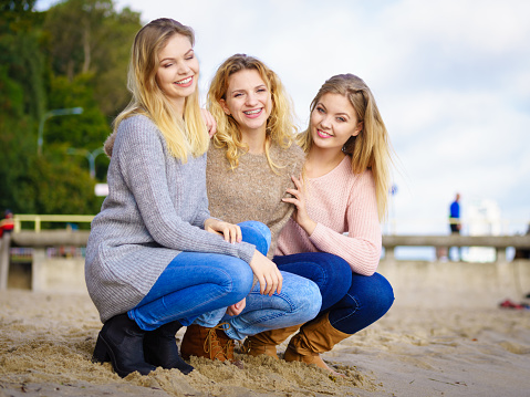 Three fashionable women wearing sweaters during warm autumnal weather spending their free time on sunny beach. Fashion models outdoor