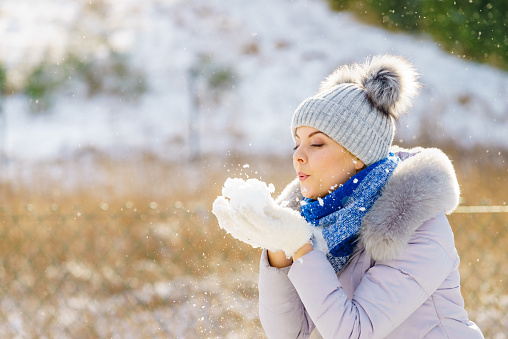 Pretty young woman blowing snow playing with it. Female having grey beanie warm hat with pompons and blue scarf.