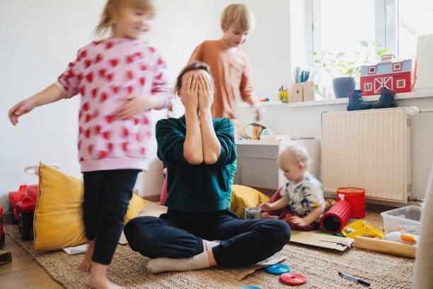 Stressed out mother sitting on floor in middle of toys while children naughty running around her at room. Woman alone burnout with kids. Family home with chaos, mess. Motion blur for speed, real life.
