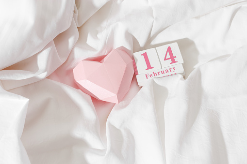 Valentine's Day concept, pink paper valentine heart and 14 february holiday date on wooden calendar in bed on on  white crumpled sheets. Minimal style flat lay photo, top view, copyspace, pastel color