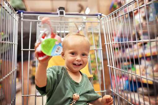 Mother with her cute daughter walking around supermarket, choosing goods and having fun. Little blonde baby girl sits in shopping trolley. Funny family weekend, happy childhood.