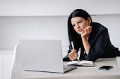 Focused brunette businesswoman working  at desk with laptop, tired looks at screen, holds pen. Pensive American woman in suit watching educational video via internet. Mockup, working people.