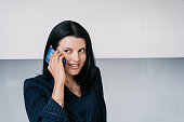 Purposeful brunette American woman talks by phone toothy smiles looks away standing indoors against wall with copy space, mockup. Businesswoman chatting.