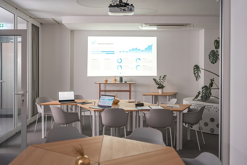Wireless technology and laptops characterize this contemporary corporate office space. A projector displays statistics on a blank white wall, devoid of people.