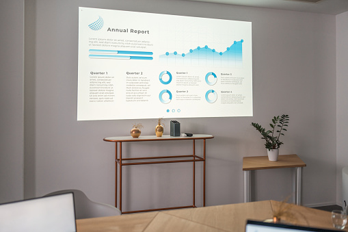 In a contemporary and well-lit corporate environment, wireless technology and laptops are prominent. A projector showcases statistics on a blank white wall, without any people present.