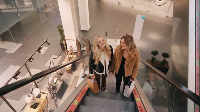 SLO MO Happy Young Female Friends in Trendy Clothes With Paper Bags Talking on Escalator in Shopping Mall