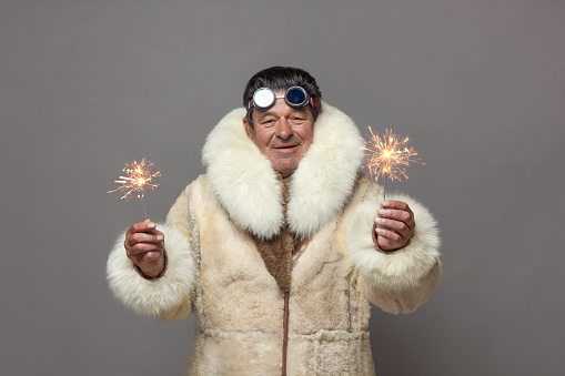 A mature man in a warm fur coat with a white fox collar and sunglasses holds a Christmas firework, a sparkler, in each hand.